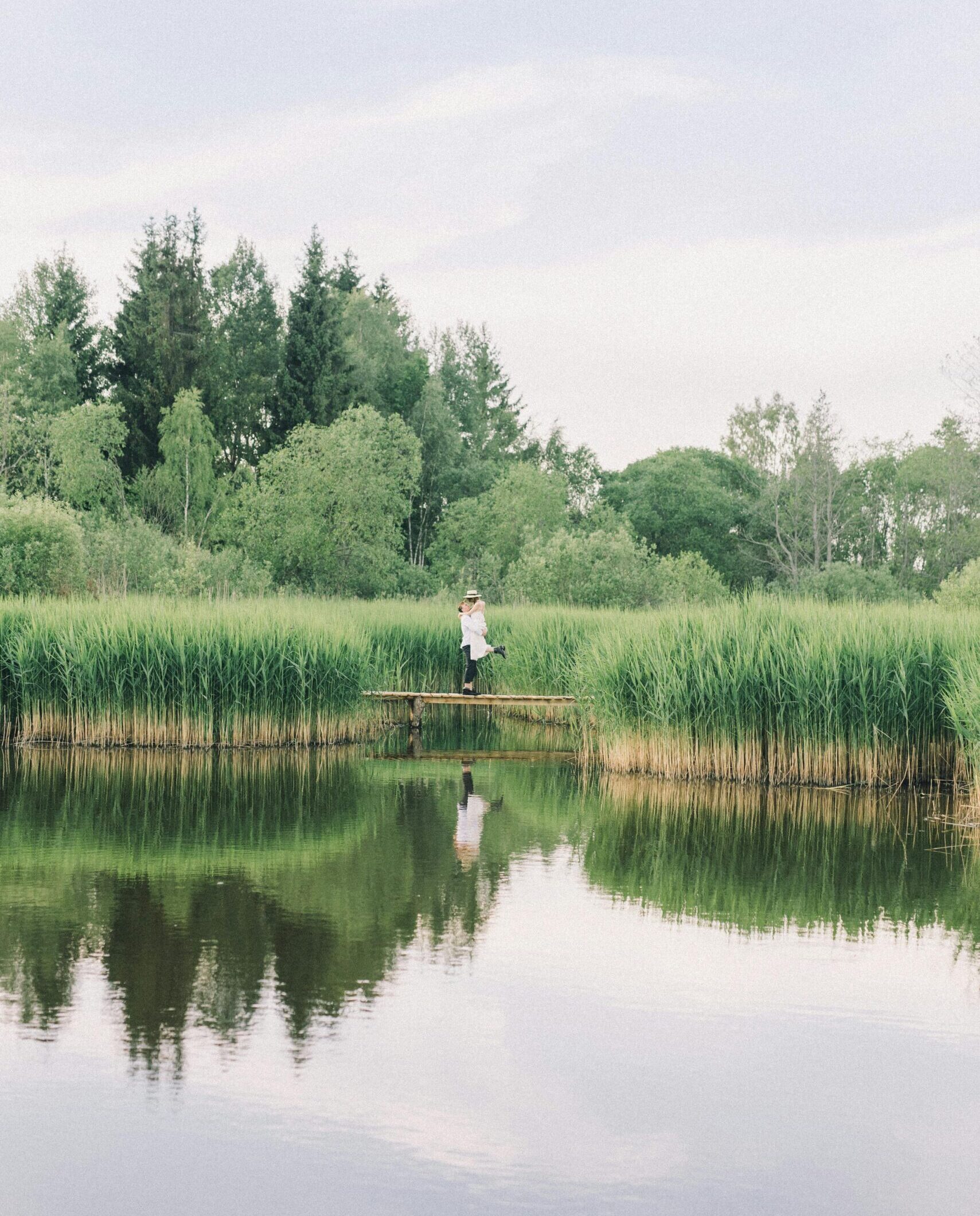 A person stands on a wooden boardwalk reflecting in a tranquil lakeside setting amidst lush reeds.
